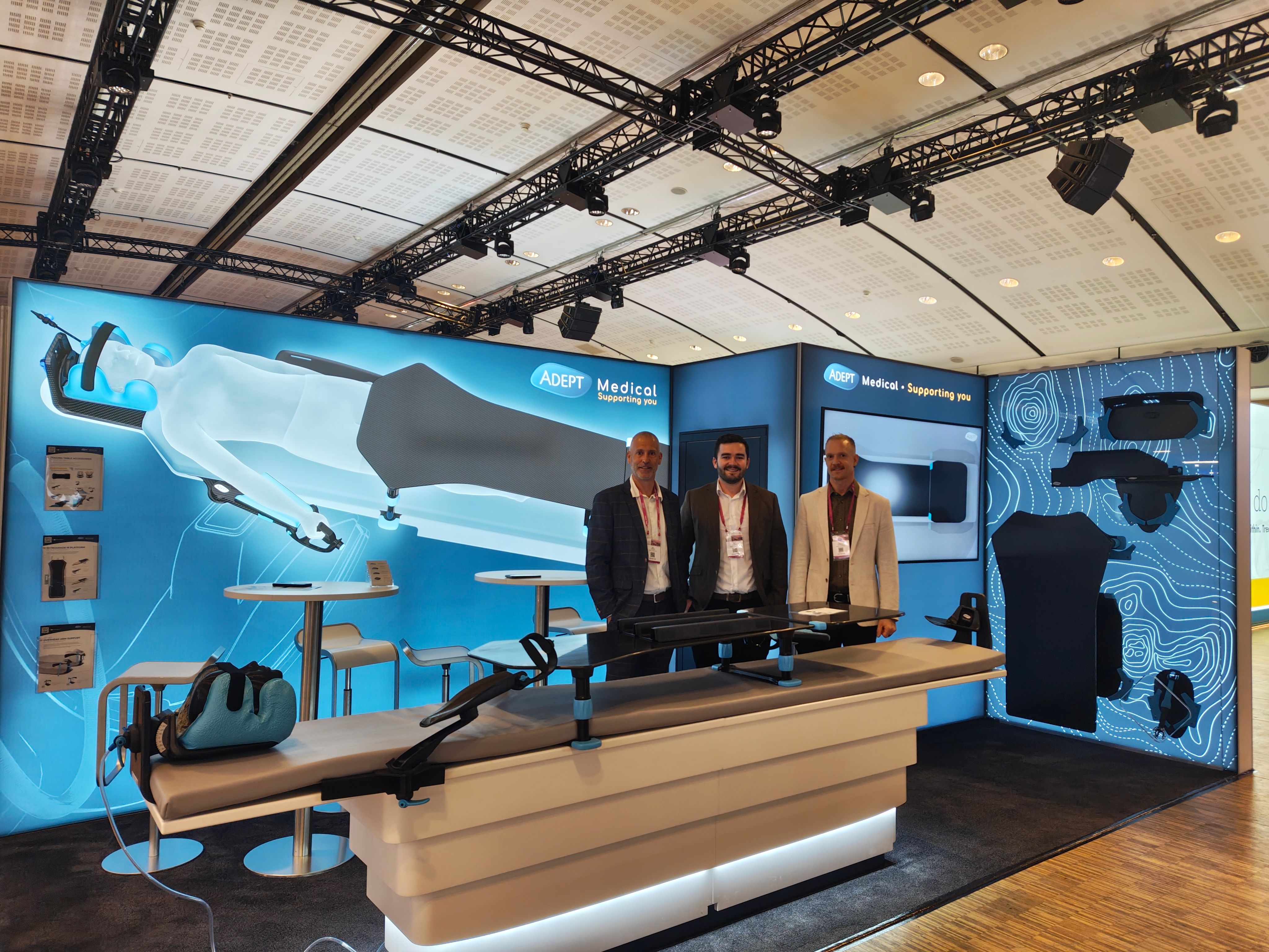 Three men stand at an ADEPT Medical trade show booth displaying medical positioning equipment and accessories.