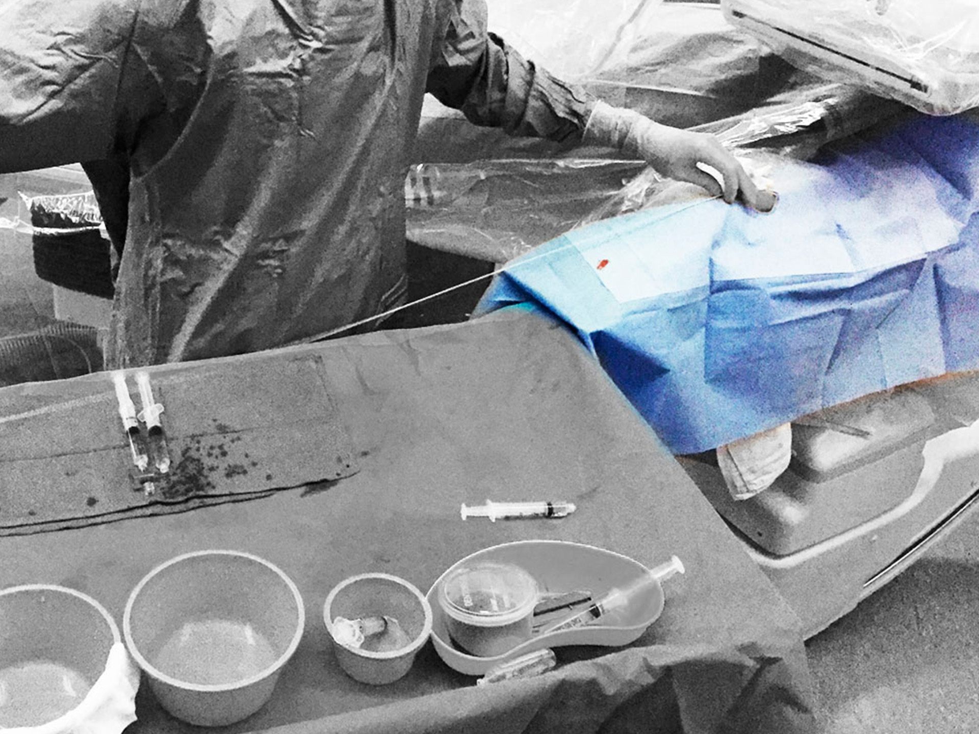 A medical professional's gloved hand rests on a blue surgical drape with a red blood spot, surrounded by grayscale medical tools.
