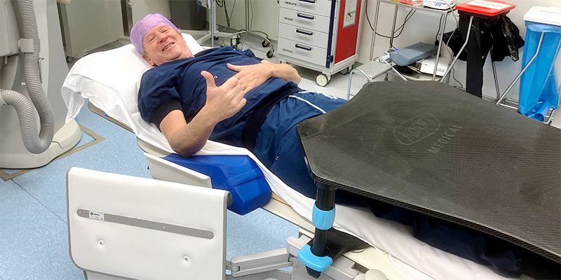 A smiling man in blue scrubs and a pink surgical cap lies on a medical bed, giving a thumbs-up, with a black medical device covering his lower body.