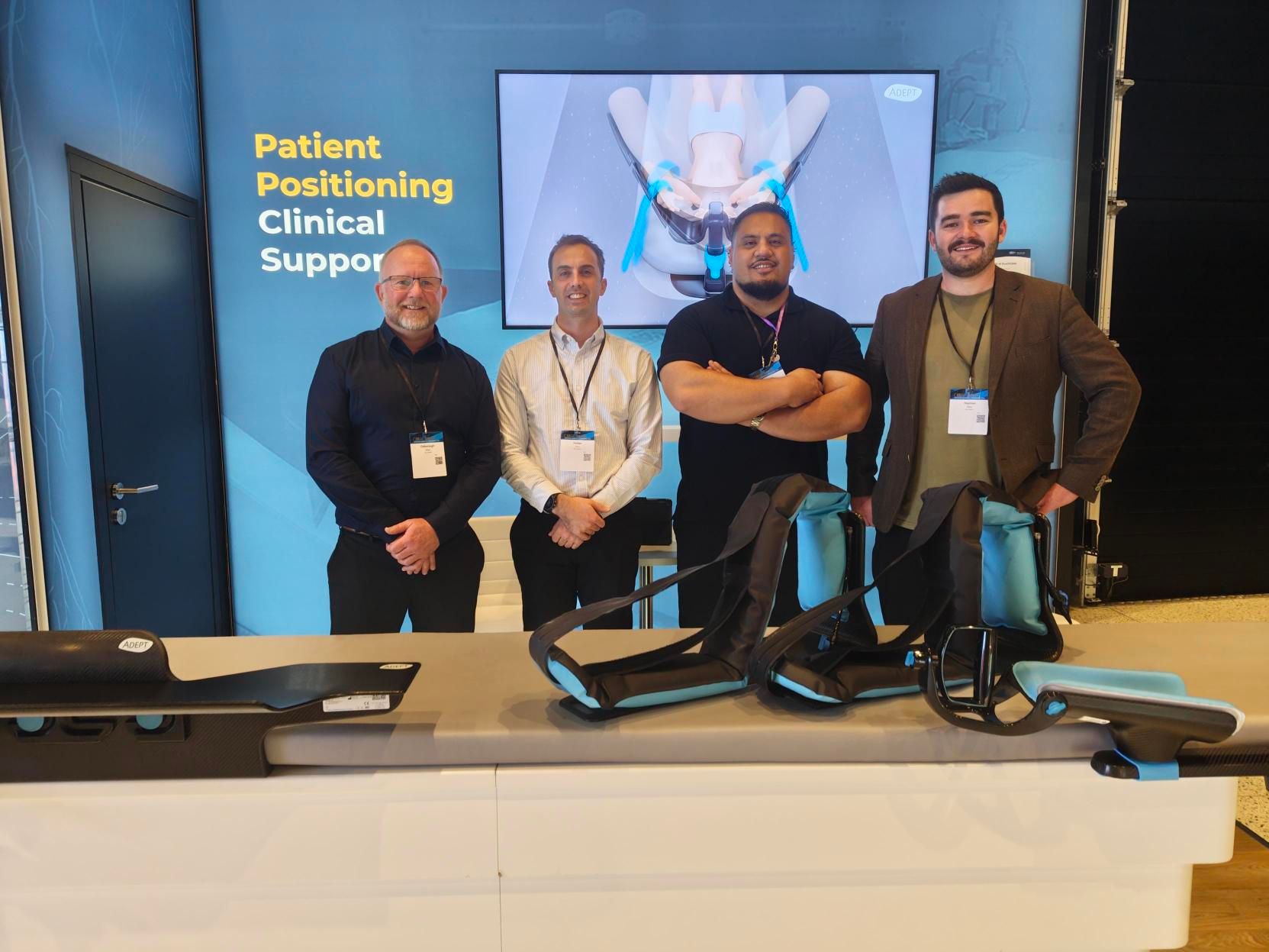 Four men stand at a medical booth displaying patient positioning devices, with "Patient Positioning Clinical Support" on the wall behind them.