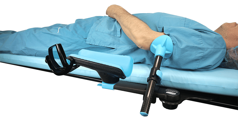 A patient in blue scrubs lying on a medical table with their arm supported by a blue and black surgical arm positioner.