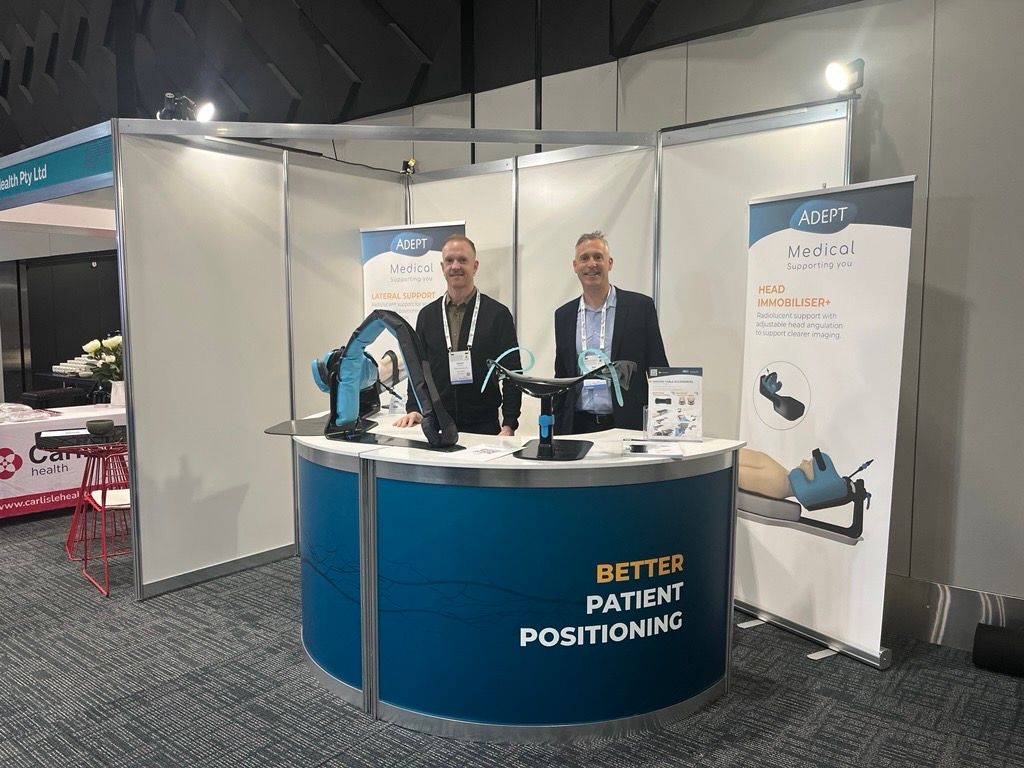 Two men stand at an Adept Medical trade show booth with patient positioning devices and banners.