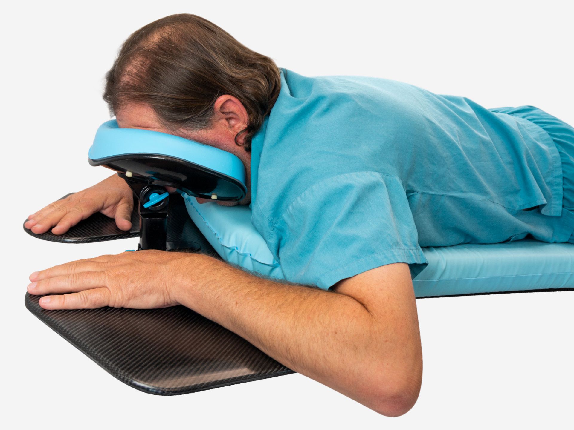 A person in blue scrubs lies face down on a table with their head in a face cradle and hands on armrests.